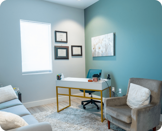 Therapy office with a desk, teal chair, gray armchair, sofa, and wall art creating a calm atmosphere.