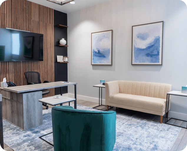 Modern therapy office with wooden accent wall, desk, cream sofa, teal chair, and framed blue artwork.